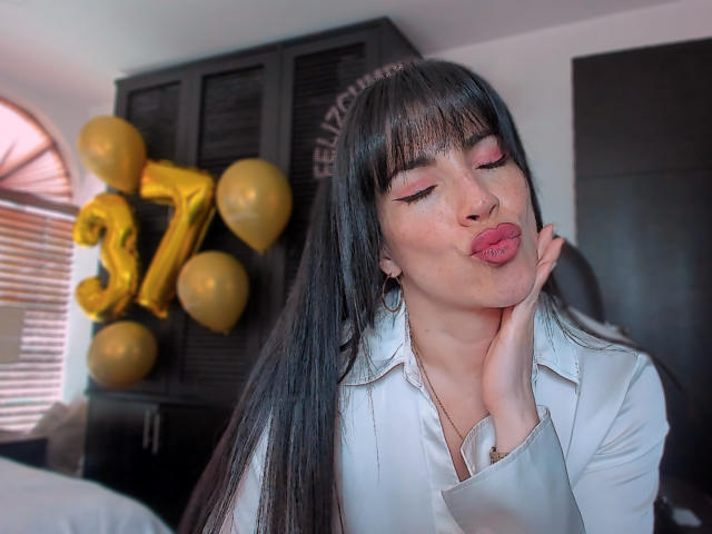 Liliysweet, con cabello negro largo y flequillo, haciendo pucheros con los ojos cerrados, con una mano en la mejilla, vestida con una camisa blanca. El fondo incluye globos dorados y muebles de madera oscura. Tensión sexual evidente..