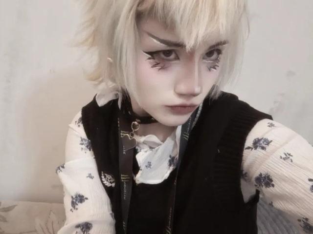 Gros plan de KatlynRenteria avec des cheveux blonds en bataille, un trait d'eye-liner noir prononcé et une peau pâle. Elle porte un gilet noir par-dessus une chemise blanche à motifs floraux, et regarde vers le bas avec une expression sérieuse. Froncement des sourcils.