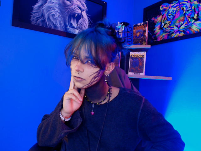 Dandii69, a slender androgynous person with blue hair and bangs, gazes seductively at the camera with a finger on their cheek, wearing a black hoodie. Neon blue lighting enhances the scene. Vibrant wall art in background.