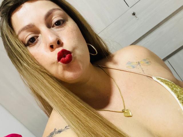 Autofoto de primer plano de SamanthaGold, una mujer de piel clara con cabello largo castaño, lápiz labial rojo, aretes de aro y un collar de oro. collar, frunciendo los labios. Ella tiene tatuajes en el hombro derecho..