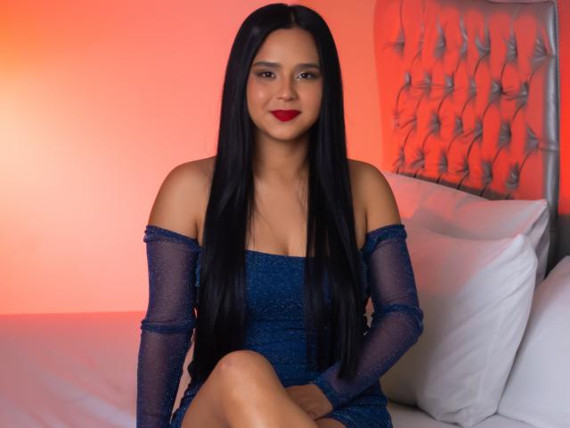 AmelyBella está sentada en una cama, vestida con un vestido azul brillante con un hombro descubierto y mangas transparentes, con lápiz labial rojo y cabello negro largo. Fondo rojo anaranjado, cabecera acolchada, almohadas blancas, ambiente sexy y seductor..