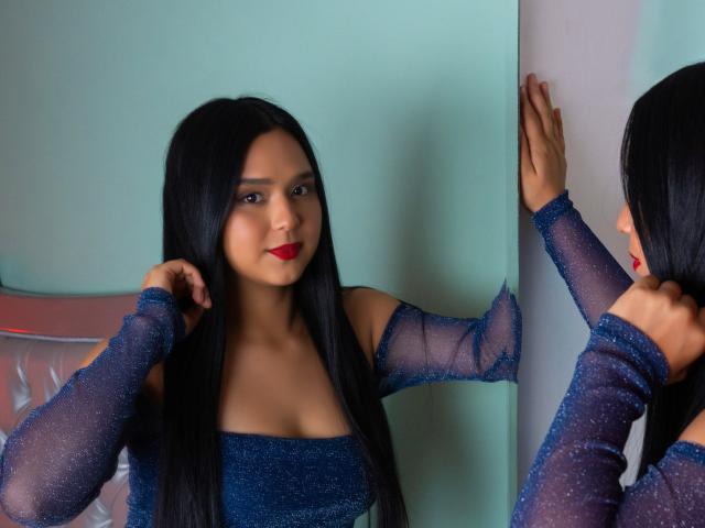 AmelyBella, een aantrekkelijke vrouw met lang zwart haar, rode lippenstift en een glinsterende blauwe jurk, staat tegen een muur, haar rechter haar hand tegen het oppervlak, haar linkerhand dicht bij haar gezicht. Reflectie.