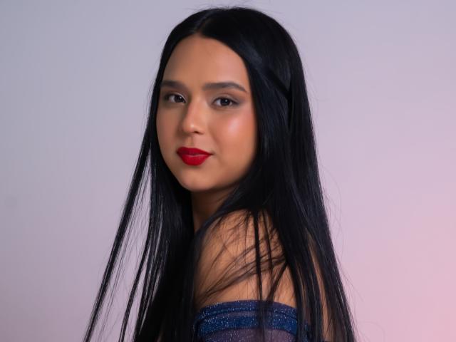 Fotografia de AmelyBella, uma mulher latina atraente com cabelos longos e pretos, batom vermelho e um vestido azul brilhante sem alças. Ela está virada ligeiramente para o lado, olhando por cima do ombro com um olhar sedutor..