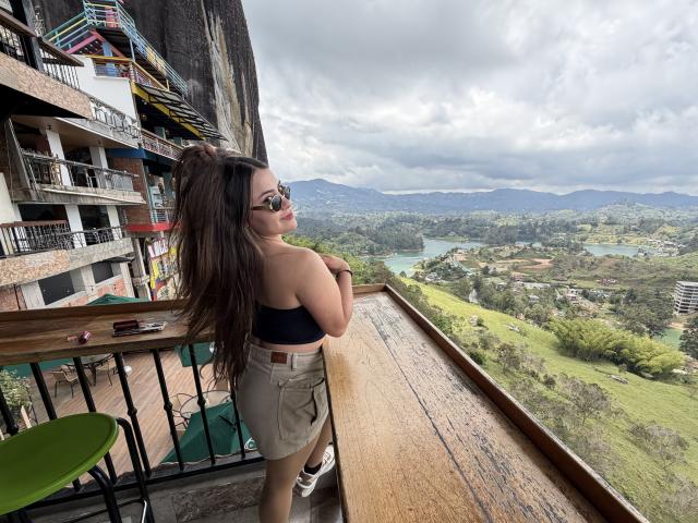 LorenzDouce with long brown hair, wearing sunglasses and a black top, stands on a balcony, looking out over a mountainous landscape. She's in beige shorts, cock not visible. Bright, cloudy sky.
