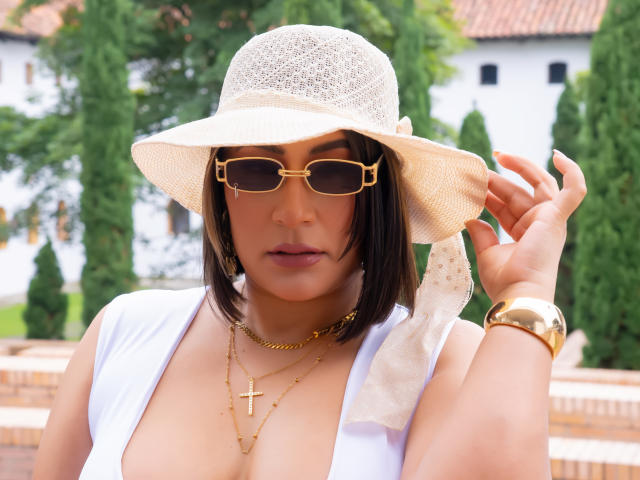 EmaFoxy, tan-skinned with black bob, wears white sunhat, gold sunglasses, gold cross necklace, white vest, holding hat. Large breasts, gold watch. Background: green trees, white house. Slight sexual tension.