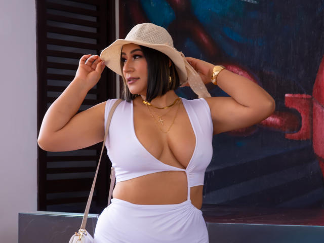 EmaFoxy, con una figura curvilínea y voluptuosa, lleva puesto un top corto blanco con un escote pronunciado y una falda a juego. Ella está ajustando un sombrero de paja, mostrando su busto grande. Joyería dorada y trenza. Pose sexy y segura..