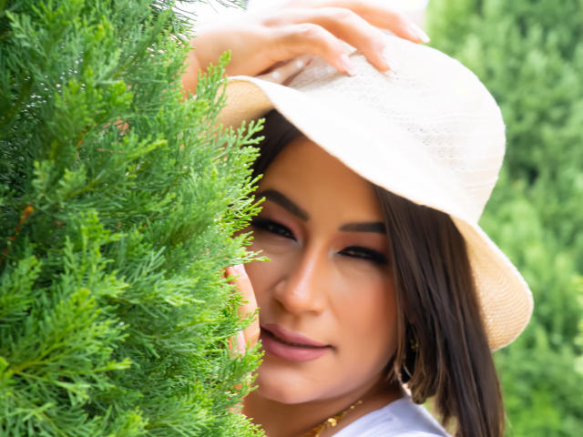 Foto em close-up de EmaFoxy, uma mulher de cabelos escuros e pele morena, usando um chapéu branco e uma camisa branca, parcialmente obscurecida por arbustos verdes e exuberantes, inclinados para frente com uma expressão sedutora.