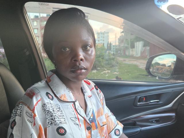 Fotografia de FineAssie sentada em um carro, olhando pela janela com uma expressão séria. Ela está vestindo uma camisa branca com padrões laranja e preto, e tem suor no rosto. A luz do sol reflete no interior do carro..