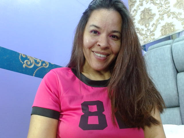 Photograph of LizethBells with long brown hair, smiling at the camera. She's wearing a bright pink shirt with black trim and a large black number "8" on the chest. She's sitting indoors.