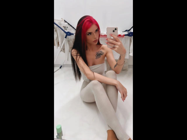 Coco1996, une femme aux longs cheveux roux et noirs, portant un haut blanc sans bretelles et un pantalon blanc, assise les jambes croisées, en train de prendre une autoportrait pris dans un miroir, tatouage visible sur la poitrine, sexy, expression confiante..