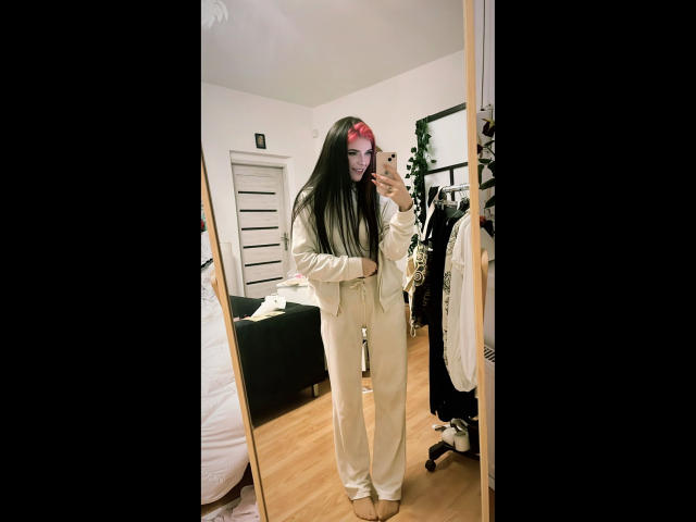 Coco1996-hot, in piedi in una camera da letto, sta scattando un selfie allo specchio. Indossava un abito bianco, ampio, con lunghi capelli neri. Lo sfondo include un letto, una cassettiera e un appendiabiti. Incredibilmente sexy..