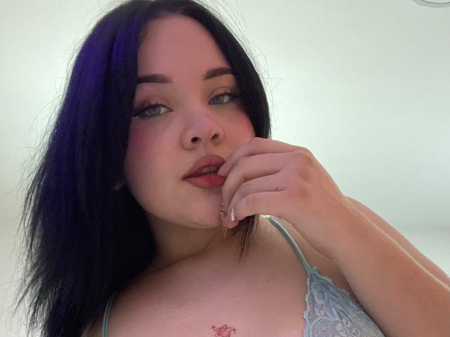 Close-up de AlayaWillow, uma mulher de pele clara com longos cabelos pretos, mordendo o dedo de forma sedutora. Ela veste uma blusa de renda azul clara, que revela uma pequena tatuagem em seu peito. Muito quente.