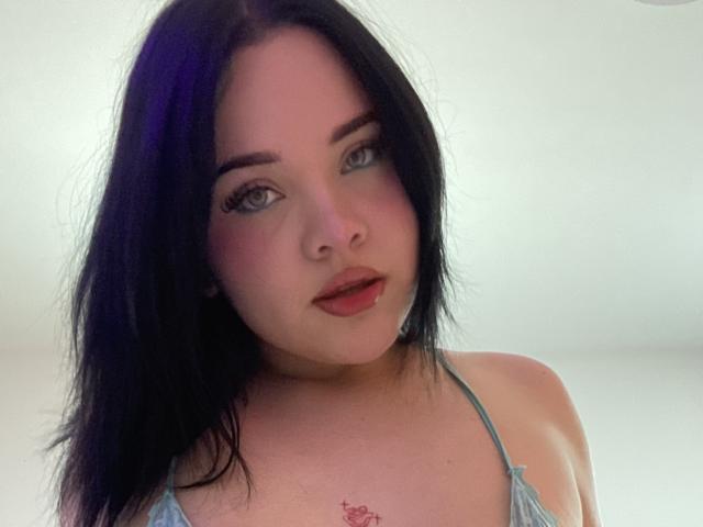 Photograph of AlayaWillow with black hair, blue eyes, and red lipstick, wearing a blue halter top, slight blush, and a small pink tattoo on her chest. Fucking sexy, direct gaze.