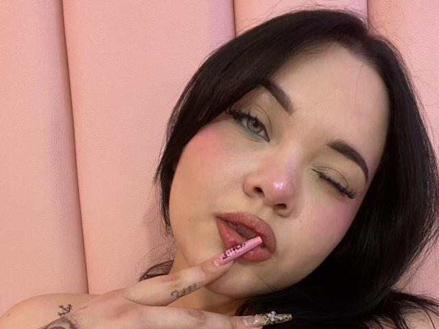 Close-up photo of AlayaWillow, winking with one eye, licking a pink lollipop with "Lollipop" written on it. She has pale skin, black hair, and green eye makeup. Background is pink.