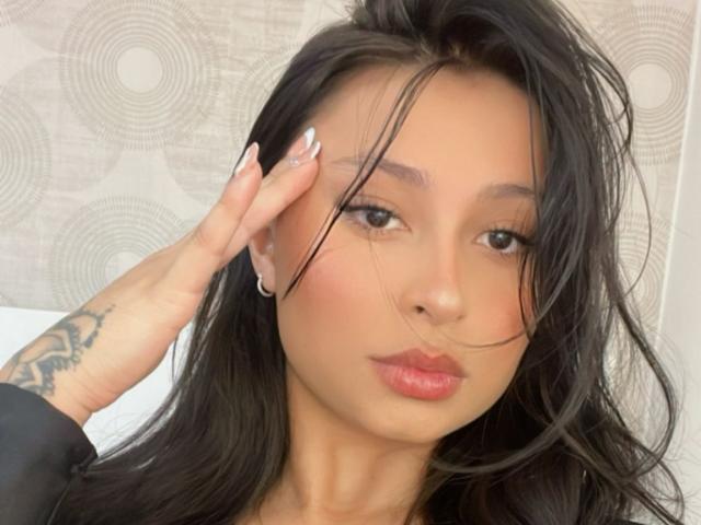 Close-up of EliaBaker, an Asian woman with long black hair, slightly messy, covering one eye with her hand. She has a tattoo on her forearm and glossy red lips. Background is white with circular patterns. Sexual tension evident in direct gaze and subtle hair play.