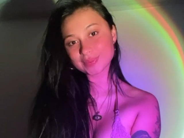 Photograph of EliaBaker, a young woman with long black hair, topless, illuminated by a neon rainbow light. Her expression is slightly playful, with visible nipples, and a heart necklace.