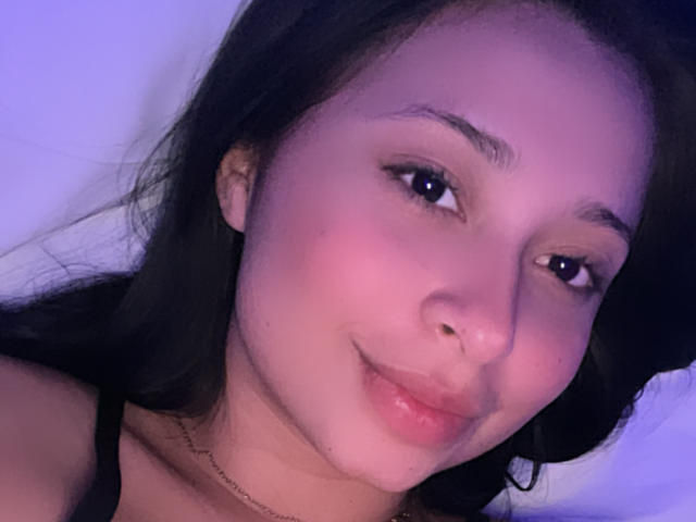 Close-up selfie of EliaBaker with soft purple lighting, dark hair, and a slight smile. She has a nose piercing, subtle makeup, and a black choker necklace. Fucking cute.