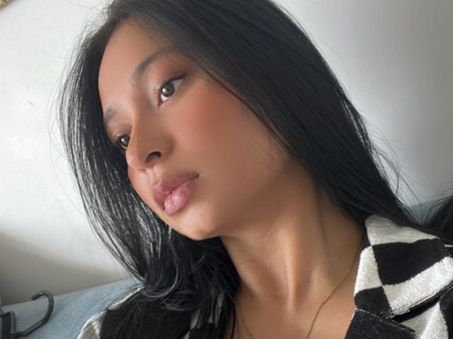 Close-up of EliaBaker, an attractive woman with long black hair, brown eyes, and full pink lips. She wears a black-and-white checkered top, slightly tilted head, looking seductively at the camera.