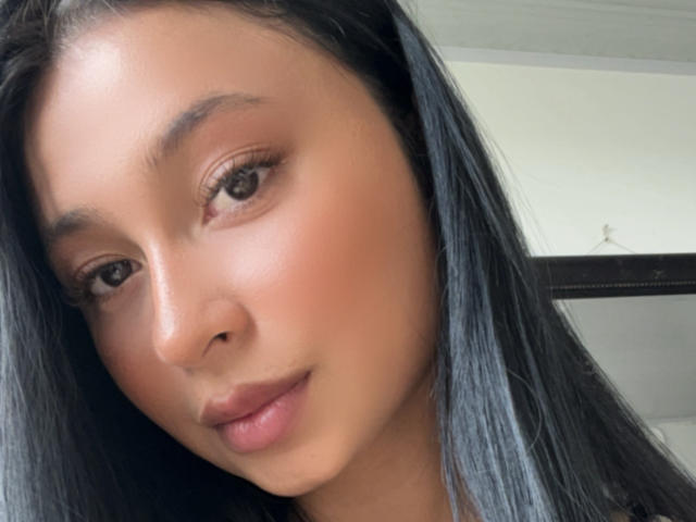 Close-up photo of EliaBaker, an Asian woman with long black hair, smooth brown skin, and full lips. She has a slight smile and direct, slightly sultry gaze. Natural light highlights her face.