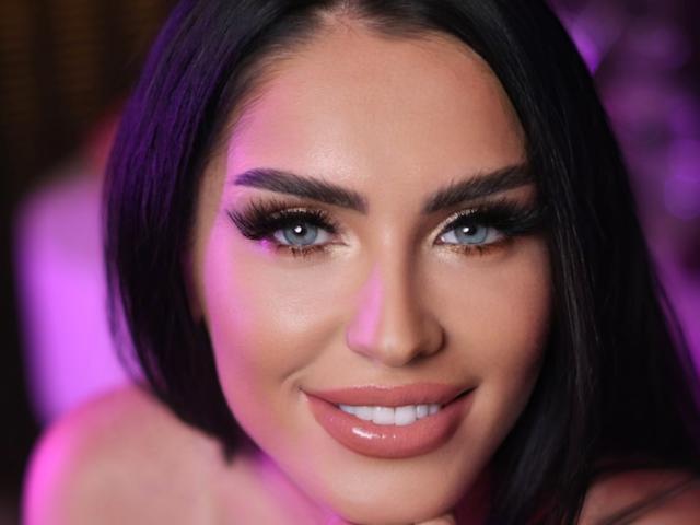 Close-up foto van RubyConner, een aantrekkelijke vrouw met donker haar, blauwe ogen en volle lippen. Ze glimlacht verleidelijk, met veel oogmake-up en roze verlichting. Haar gezicht heeft een licht glanzende uitstraling..
