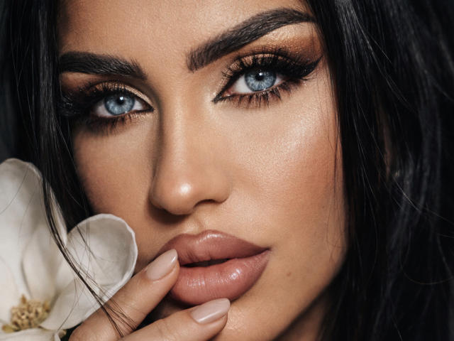 Close-up of RubyConner's face, intense blue eyes, thick dark eyebrows, glossy pink lips, black hair, holding a white flower near her face, with a sexy, seductive look, prominent cheekbones.