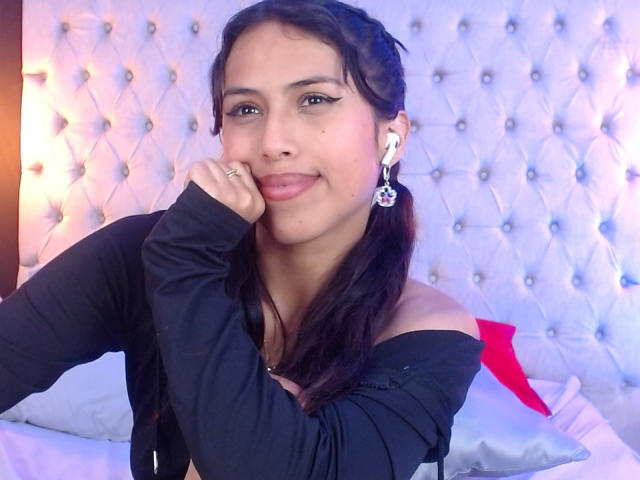 MyaaTyler with long dark hair, black off-shoulder top, hand on cheek, smiling. Tufted white headboard, red pillow. Earrings, soft lighting, casual sexy vibe. Fingernail ring visible.