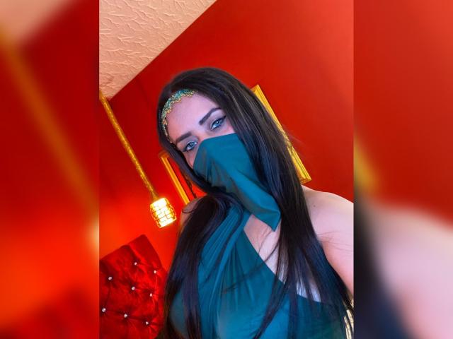 Photographie d'Anissa Ali avec de longs cheveux noirs, portant un masque noir transparent qui couvre sa bouche, et un serre-tête scintillant. Des murs rouges, un éclairage rouge, et un coussin rouge flou en arrière-plan. Tension sexuelle.