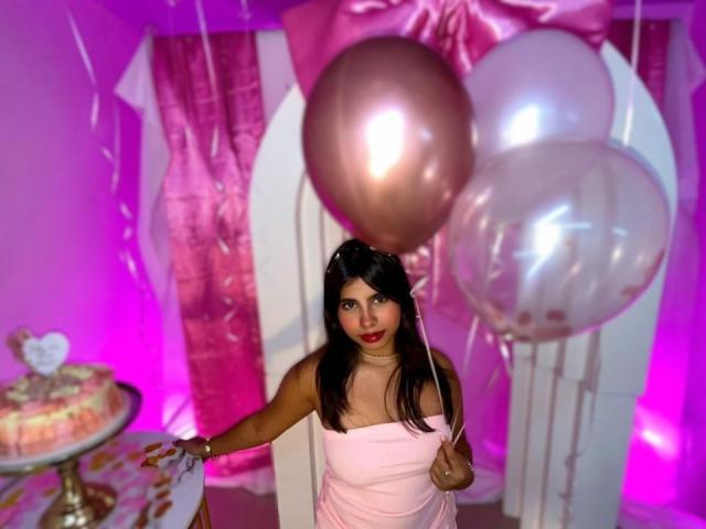 CelesteMorel, brown-skinned woman with black hair, wearing a white strapless dress, standing in front of pink and white balloons, holding one, neon pink and purple lighting, cake in background.