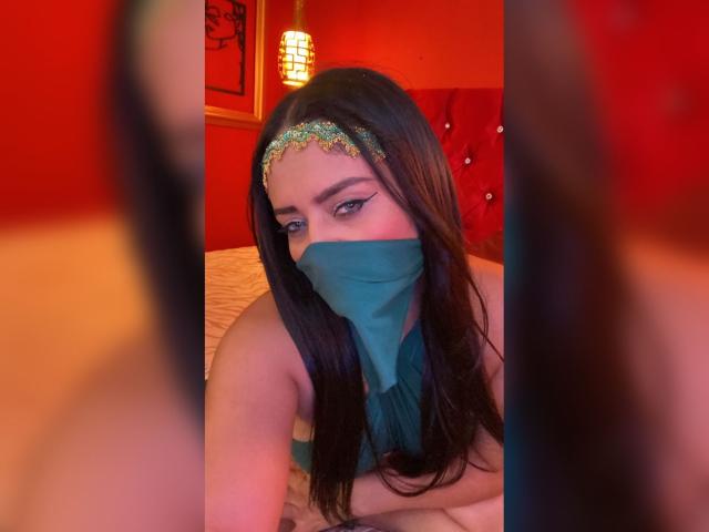 Anissa Ali, una donna con lunghi capelli neri, che indossa una fascia scintillante e una maschera blu, guarda seducentemente la telecamera. Occhi azzurri.
la luce rossa illumina la stanza, creando una forte tensione sessuale..