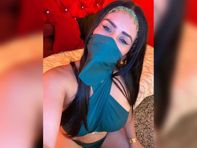 AnissaAli, a Latina woman with long black hair, wears a black mask and matching bikini, adorned with a gold headband. Her large breasts are prominent. Red and beige background. Seductive pose.