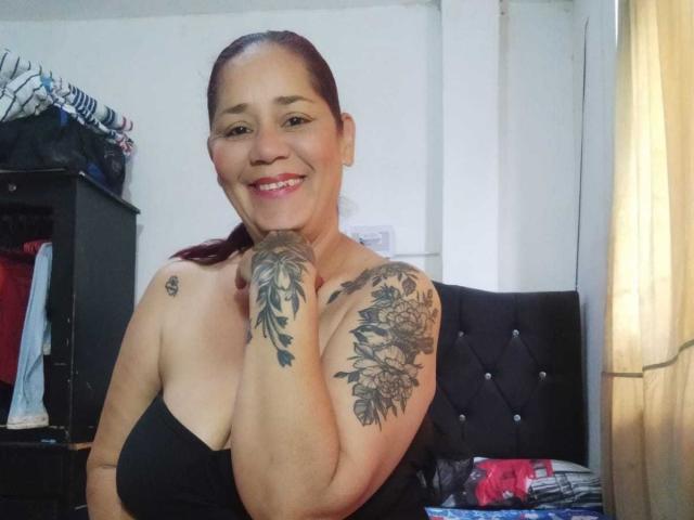 DeboraMilf69, smiling with dark hair in a ponytail, black strapless top, large floral tattoos on right shoulder and upper arm, standing in a room with a black headboard and soft light.