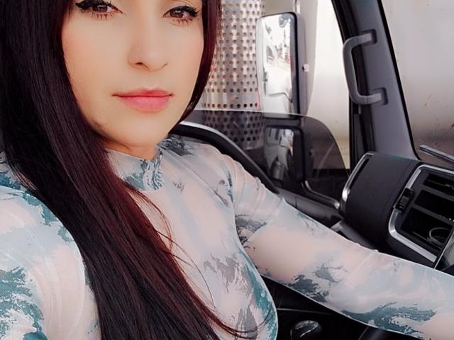 Close-up de LuluFrancaise, uma mulher de cabelos longos e pretos, vestindo uma blusa branca e azul com estampas, sentada em um carro, com a mão no volante, com um leve sorriso, os olhos fixos à frente.