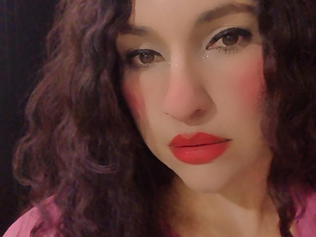 Nahaufnahme von LuluFrancaise mit lockigem, braunem Haar, rotem Lippenstift und rosa Rouge. Ihre braunen Augen glänzen leicht. Ihr Ausdruck ist leicht verführerisch. Der Hintergrund ist dunkel und unscharf..