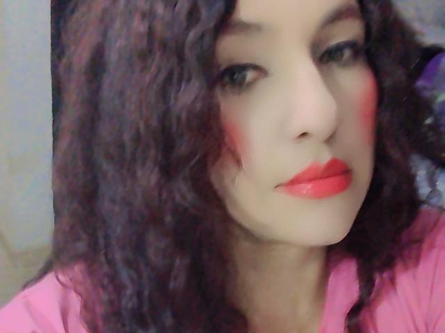 Primer plano de LuluFrancaise con cabello castaño rizado, piel clara y lápiz labial rojo brillante. Ella lleva una blusa rosa, con una mancha roja en su mejilla derecha. Increíblemente atractivo/a..