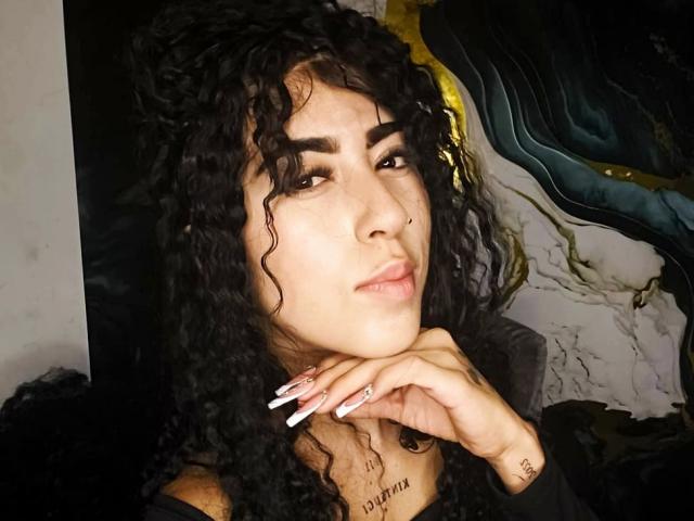 Fotografía de primer plano de PeytonMorgan, una mujer atractiva con cabello negro rizado, piel marrón clara y labios carnosos, que mira de forma seductora. frente a la cámara. Su mano, con uñas largas y blancas, descansa sobre su barbilla. El fondo presenta patrones abstractos en negro, amarillo y blanco. Ella tiene un tatuaje en el cuello que dice "SUEÑA EN GRANDE".".