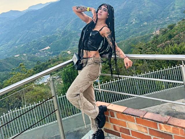 PeytonMorgan, black-haired, tattooed, wearing black crop top and beige pants, leaning on railing, mountain background. Long braids, confident pose, mountains in distance, metal railing, brick ledge. Fucking sexy.