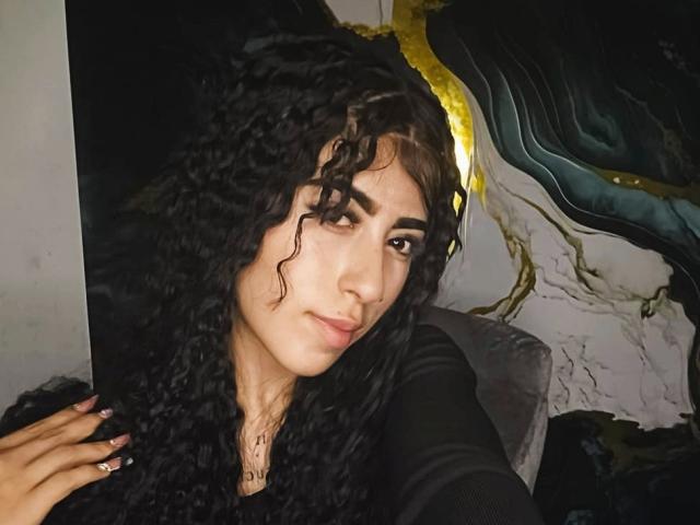 Close-up of PeytonMorgan with curly black hair, light brown skin, and dark eyebrows, wearing a black top, leaning against a dark abstract wall with gold and blue accents. Sexy, direct gaze.