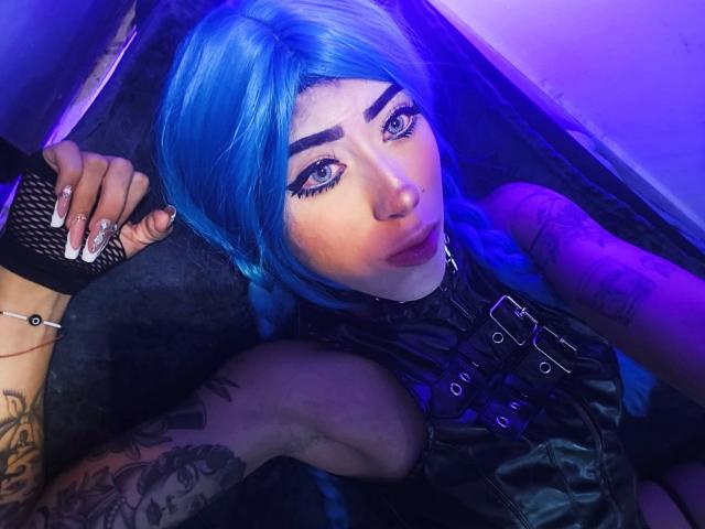 Close-up van PeytonMorgan met felblond haar, intense blauwe ogen en donkere make-up. Een zwarte leren top aan, hand rustend op het hoofd, blauwe verlichting, tatoeage op de arm, verleidelijke uitdrukking..