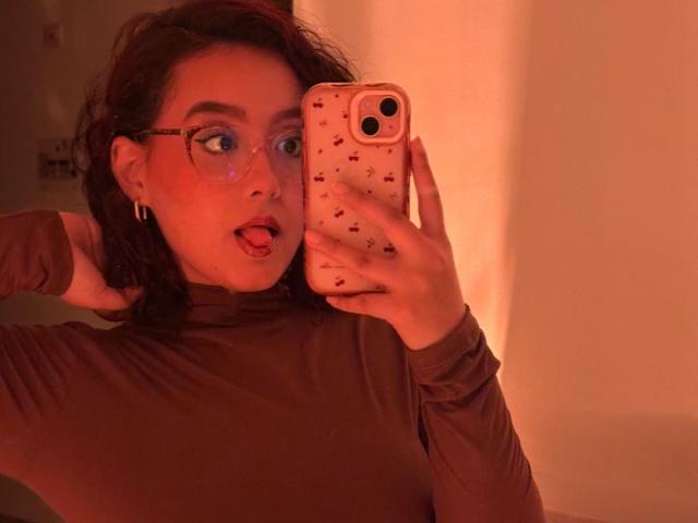 Foto van Sophia Browm, met rode accenten in de belichting, die een spiegel-selfie maakt met een telefoon. Ze heeft donker haar, een bril en een verraste uitdrukking, en draagt een bruine coltrui. Haar tong hangt buiten..