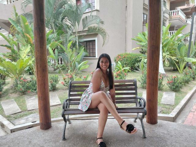 PauletteLoveX, sexy woman with tan skin and black hair, sitting on a black bench, legs crossed, wearing a white floral dress, black high heels, outdoor garden, tropical plants, wooden pillars.