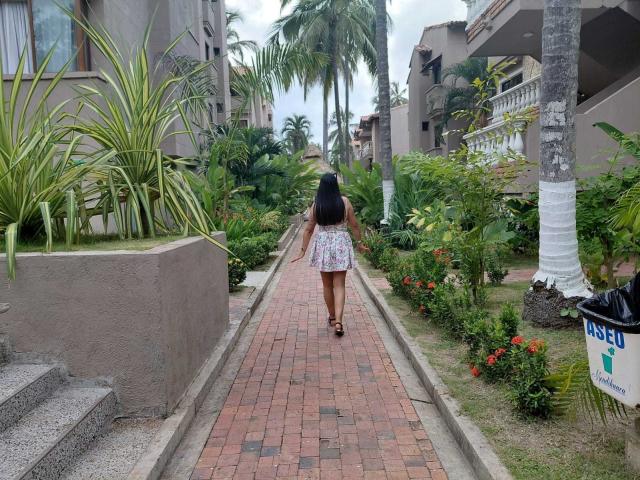 PauletteLoveX, seen from behind, walks down a red-brick path between lush green plants and palm trees, wearing a white skirt, black top, and sandals. Fucking tropical vibe, buildings in background.