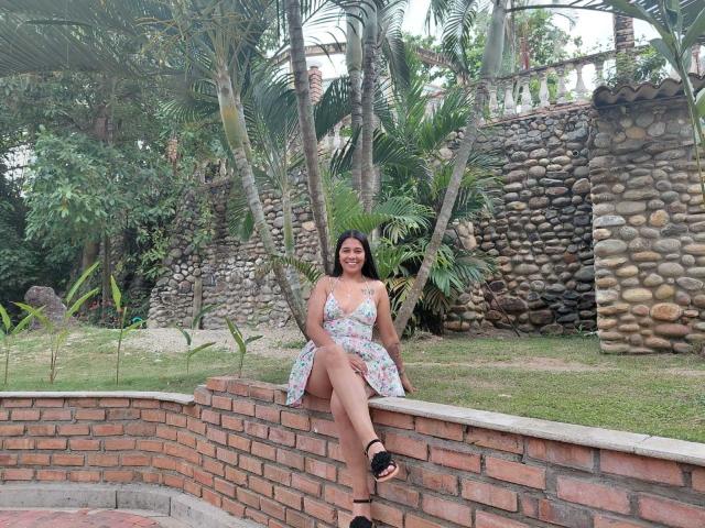 PauletteLoveX, una mujer de curvas con senos de tamaño medio, está sentada en un muro de ladrillos en un jardín tropical. Ella lleva un vestido blanco con estampado floral y tacones negros, con las piernas separadas, mostrando tensión..