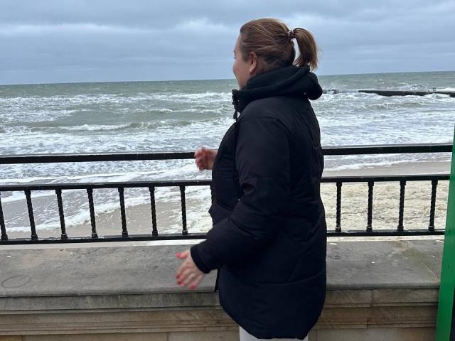 Lara Harper, gekleed in een zwarte, dikke jas, staat bij een metalen hekwerk en kijkt naar de ruwe zee. Haar handen rusten op de leuning, en ze straalt een gespannen, nadenkende sfeer uit. Bewolkte lucht.