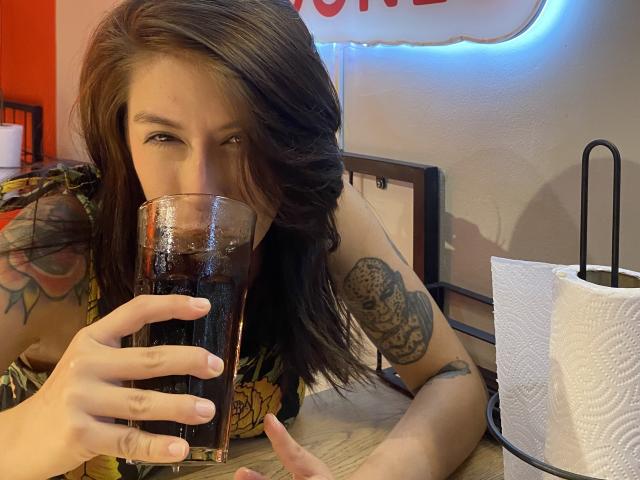 Isabella Palacio, con i suoi lunghi capelli scuri e i tatuaggi sulle braccia, si sporge in avanti e beve un liquido scuro da un bicchiere. Mano destra appoggiata sul tavolo, rotolo di carta bianca a destra. Atmosfera rilassata, tensione sessuale..