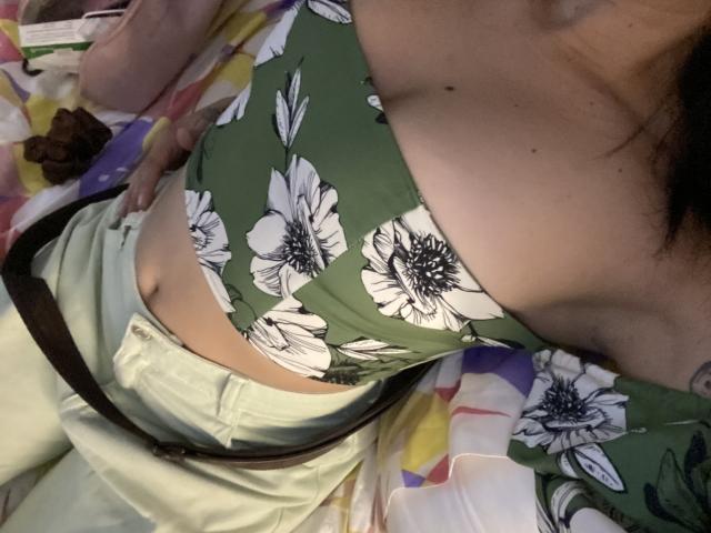 Foto em close-up de IsabellaPalacio deitada em uma cama, vestindo uma blusa verde com estampas florais que revela bastante o colo, e branca calças com um cinto preto. A pele dela é bronzeada. O tecido é justo 
.
