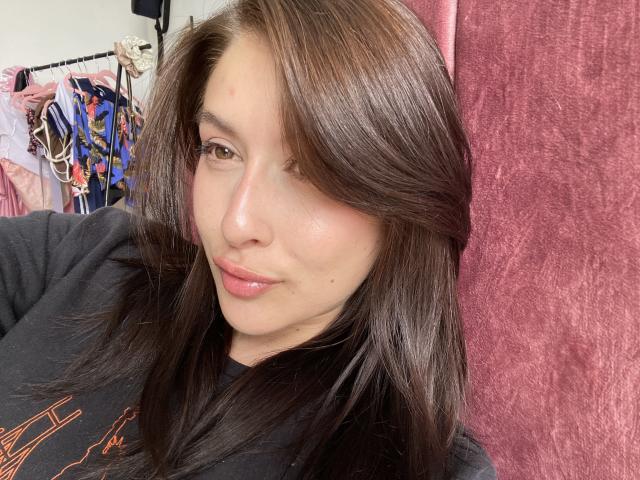 Close-up van Isabella Palacio, een vrouw met bruin haar en glanzende lippen, een zwarte top, die lichtjes naar rechts leunt. De achtergrond toont kleurrijke kleding en een rode, gestructureerde muur. Er is een lichte seksuele spanning in de blik..