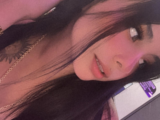 Close-up of GraceClarke, a young woman with long black hair, pink highlights, and a sun tattoo on her neck. She has a nose ring and wears a gold chain. Fucking sexy expression.