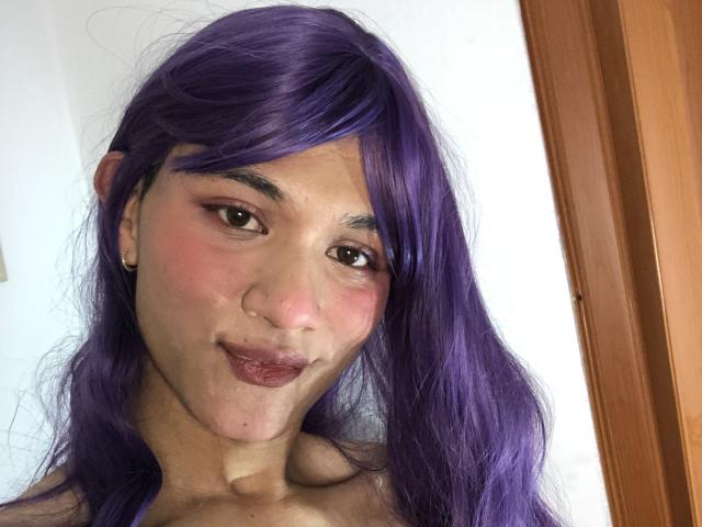 Foto ravvicinata di Holyshweet69 con capelli viola, pelle chiara e guance rosate, che guarda direttamente l'obiettivo. un leggero sorriso.
le loro labbra sono piene e leggermente carnose. Cornice in legno a destra 
.