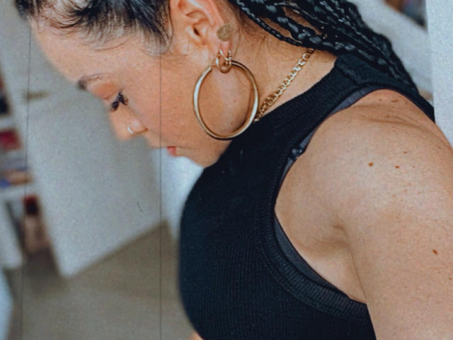 Foto ravvicinata di TrinityPrestige con trecce scure, grandi orecchini a cerchio dorati e una canotta nera a coste. Il profilo laterale di TrinityPrestige mostra uno sguardo leggermente rivolto verso il basso, che mette in risalto la sua figura sinuosa..