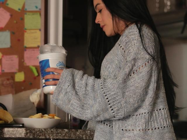TrinityPrestige with long black hair, wearing a textured gray sweater, holds a blue and white coffee cup, standing beside a countertop with a bowl of cookies. Sexual tension, casual setting.